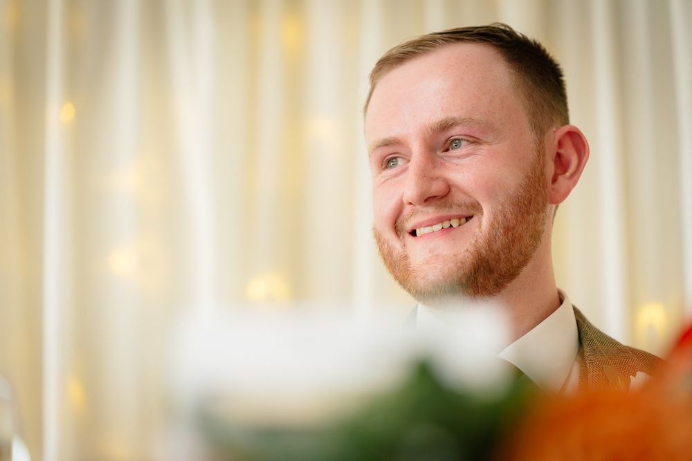 A person in a blue suit and tie smiles warmly against a soft-focus background with warm lighting.