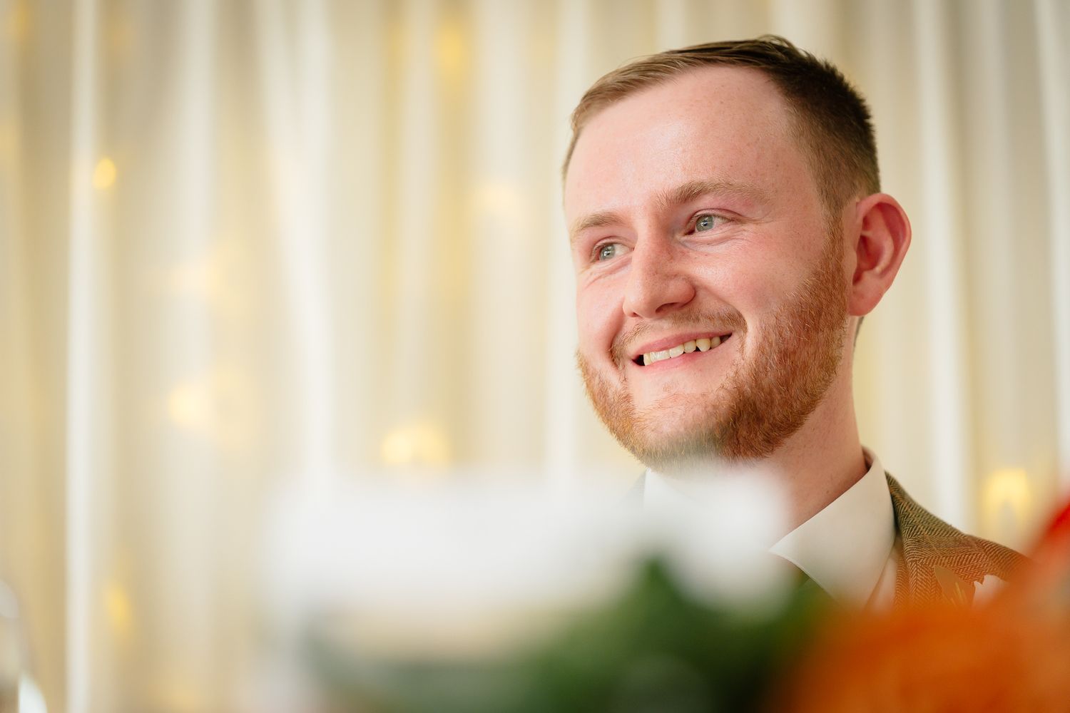 A person in a blue suit and tie smiles warmly against a soft-focus background with warm lighting.