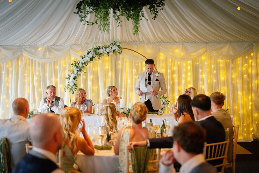 Wedding reception guests enjoy a festive atmosphere under a white draped tent ceiling with twinkling lights and greenery.