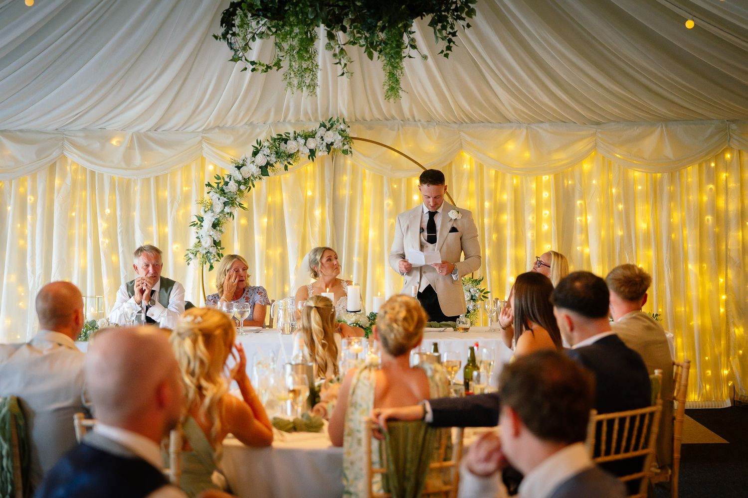 Wedding reception guests enjoy a festive atmosphere under a white draped tent ceiling with twinkling lights and greenery.