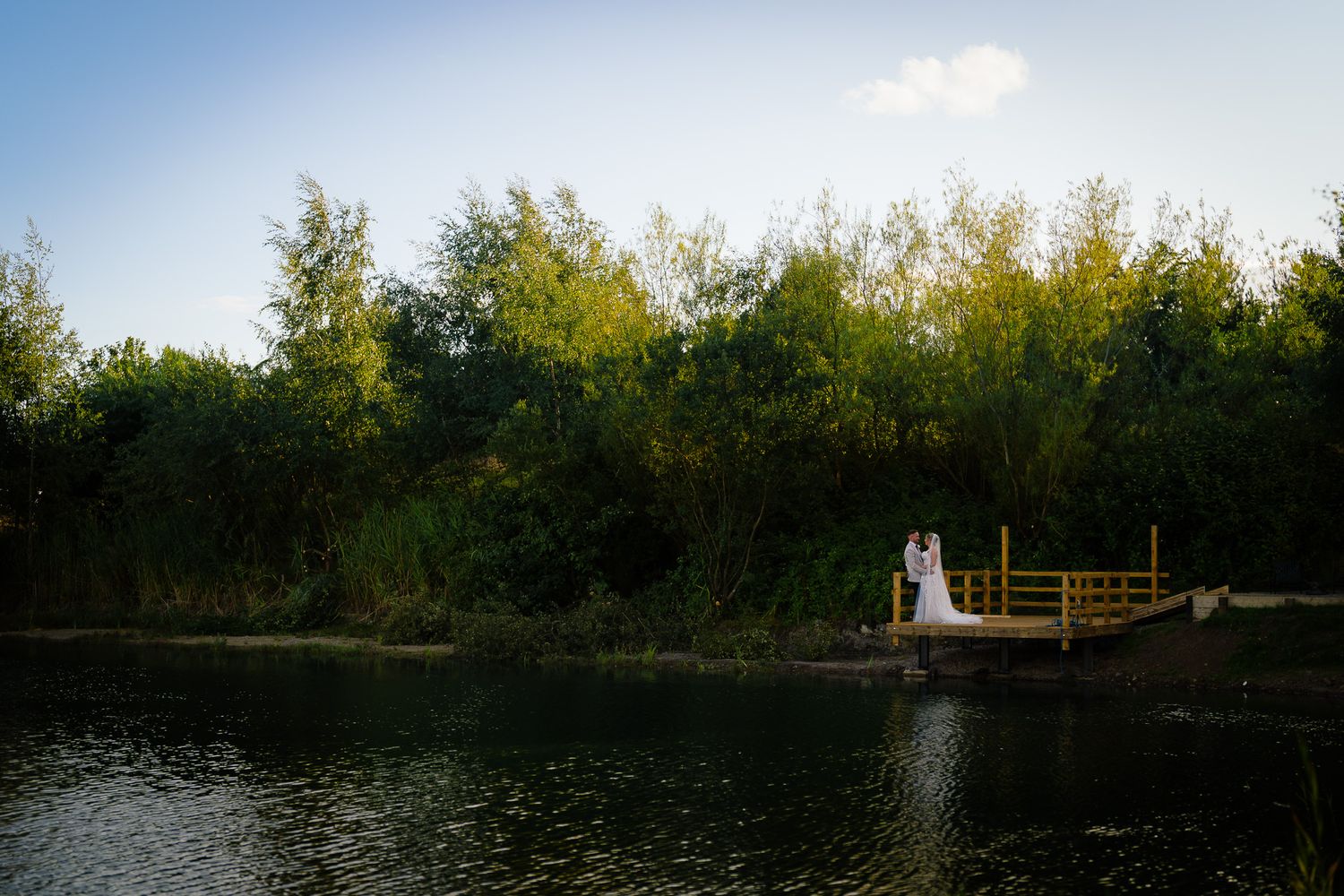 A scenic wooden bridge stretches across a tranquil pond surrounded by autumn trees and blue sky.