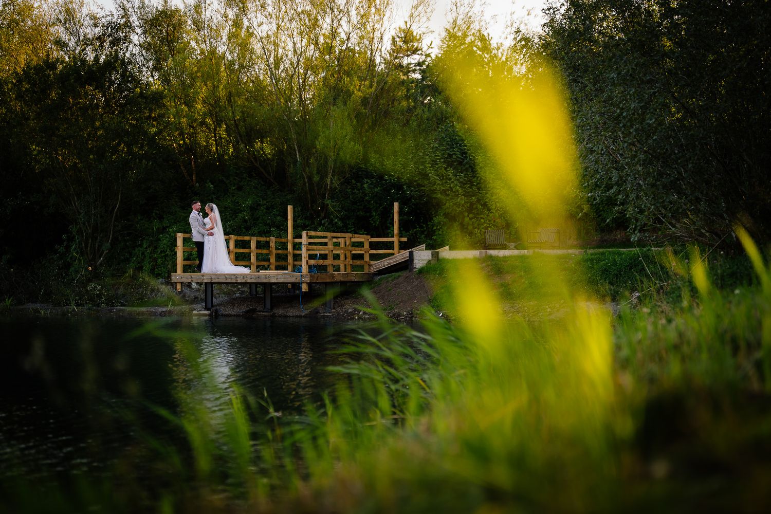 A couple stands together on a rustic wooden bridge surrounded by lush greenery at sunset.