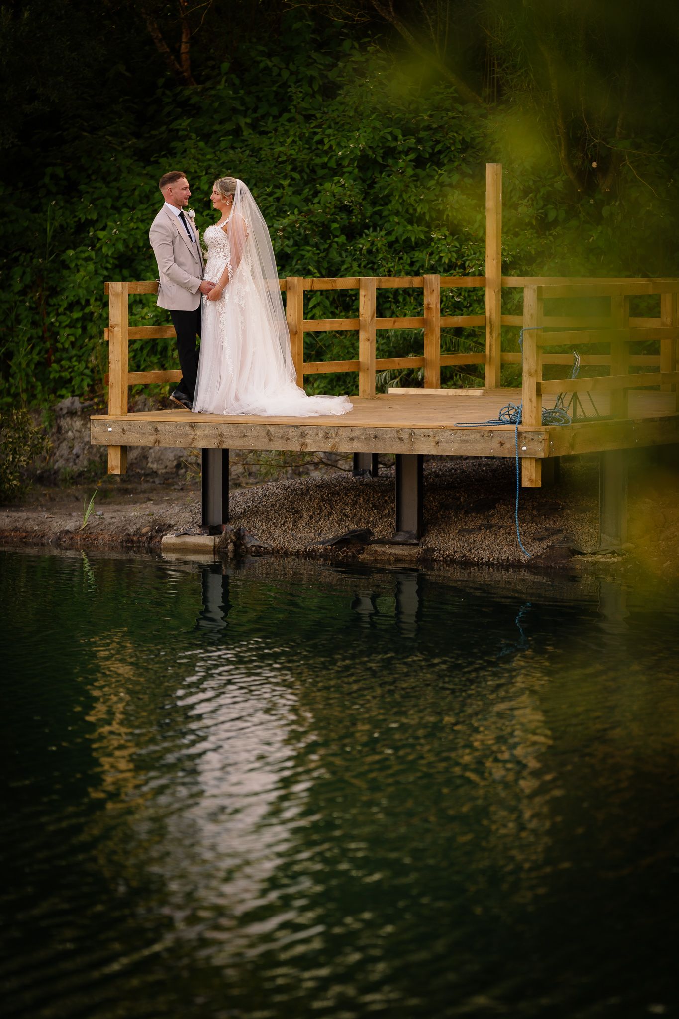 A couple in wedding attire stands on a wooden dock by a tranquil lake during sunset creating romantic silhouettes.