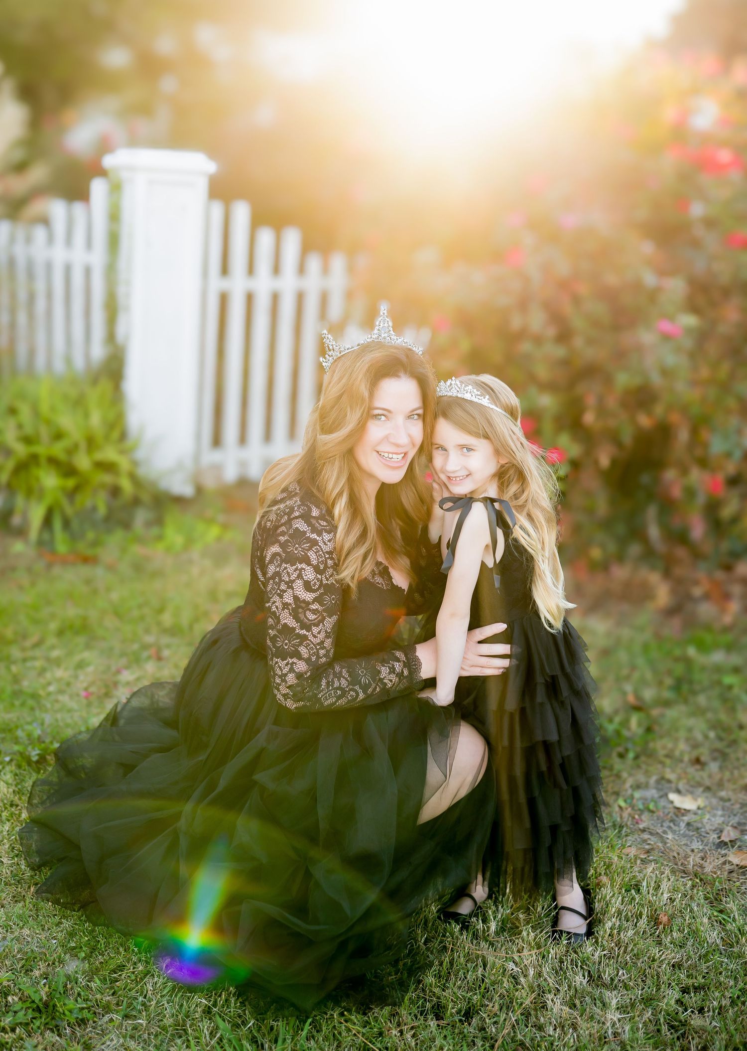 A mother and daughter wearing matching black dresses pose together in a garden with a white picket fence at sunset.