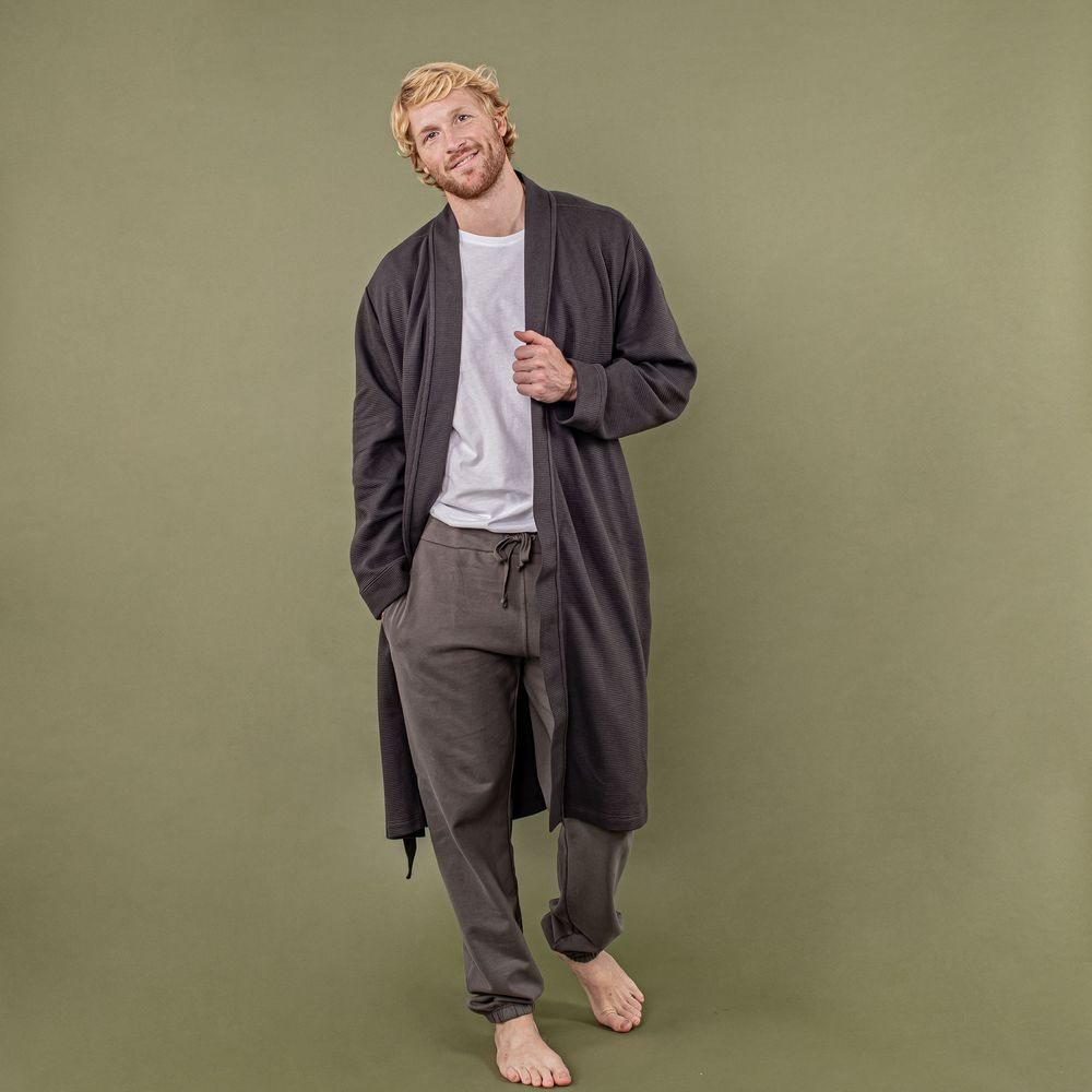 Person wearing a long dark cardigan and gray pants stands barefoot against an olive green background.