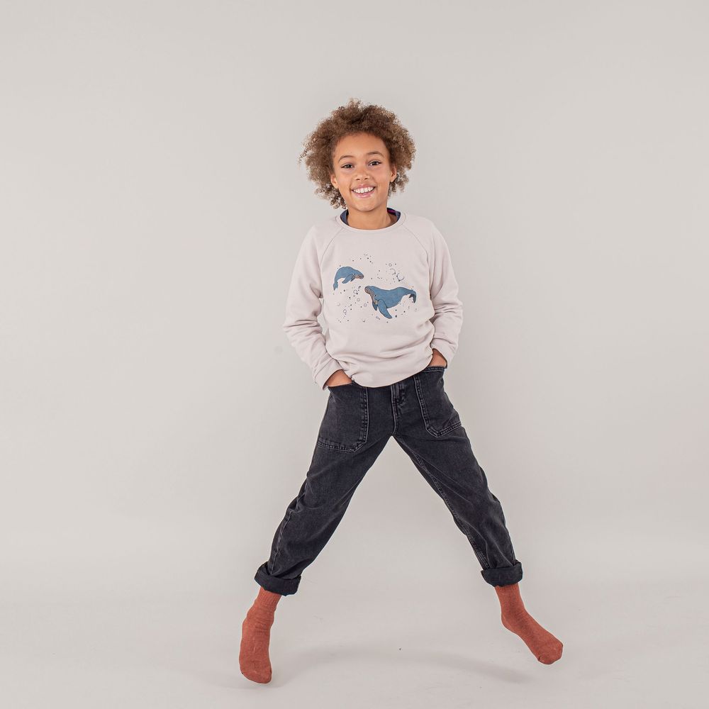 Young child wearing horse graphic sweater and jeans jumps playfully in brown socks against gray background.