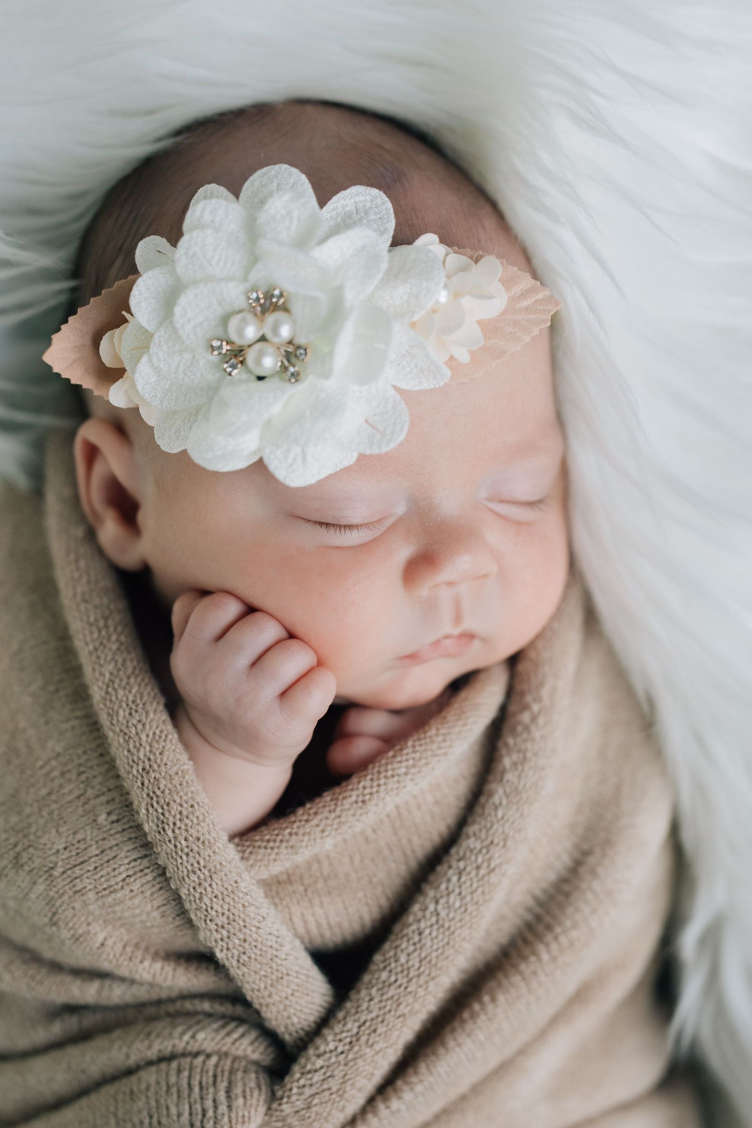 Sleeping newborn baby girl wrapped in beige knit with a white flower headband.