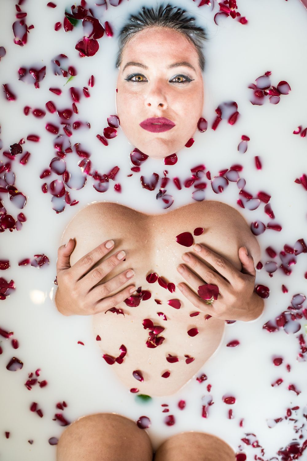 Artistic beauty portrait with scattered red rose petals against white background in milk bath style photography.