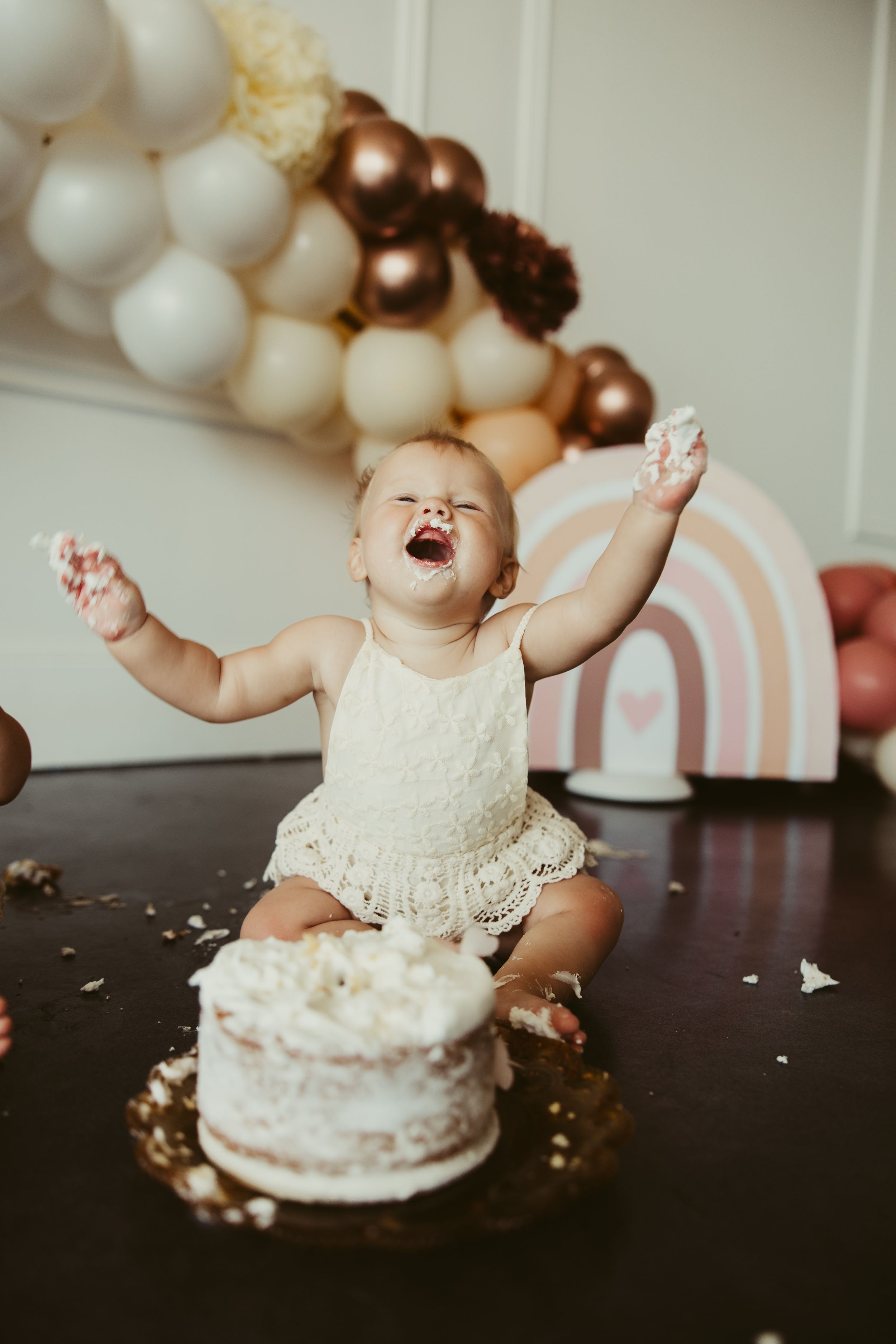 Joyful baby girl raises arms with cake-covered hands during first birthday cake smash session.