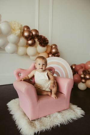 Cute baby girl sitting in pink velvet throne chair surrounded by elegant balloon garland backdrop.