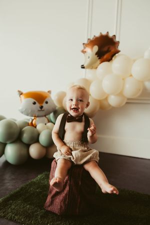 Happy baby boy in suspenders and bow tie laughing at woodland themed first birthday cake smash.