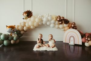 Twin babies sit on a white rug surrounded by a boho balloon garland with woodland animal decorations and a pastel rainbow.