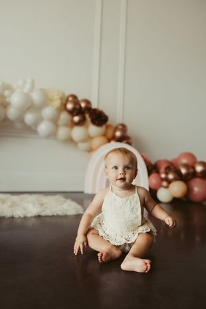 Adorable baby girl in white lace romper sits on dark floor in front of boho balloon garland and pastel rainbow decor.