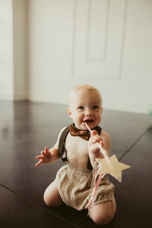 Happy baby boy in suspenders and bow tie sits on dark floor chewing a star wand at a first birthday celebration.
