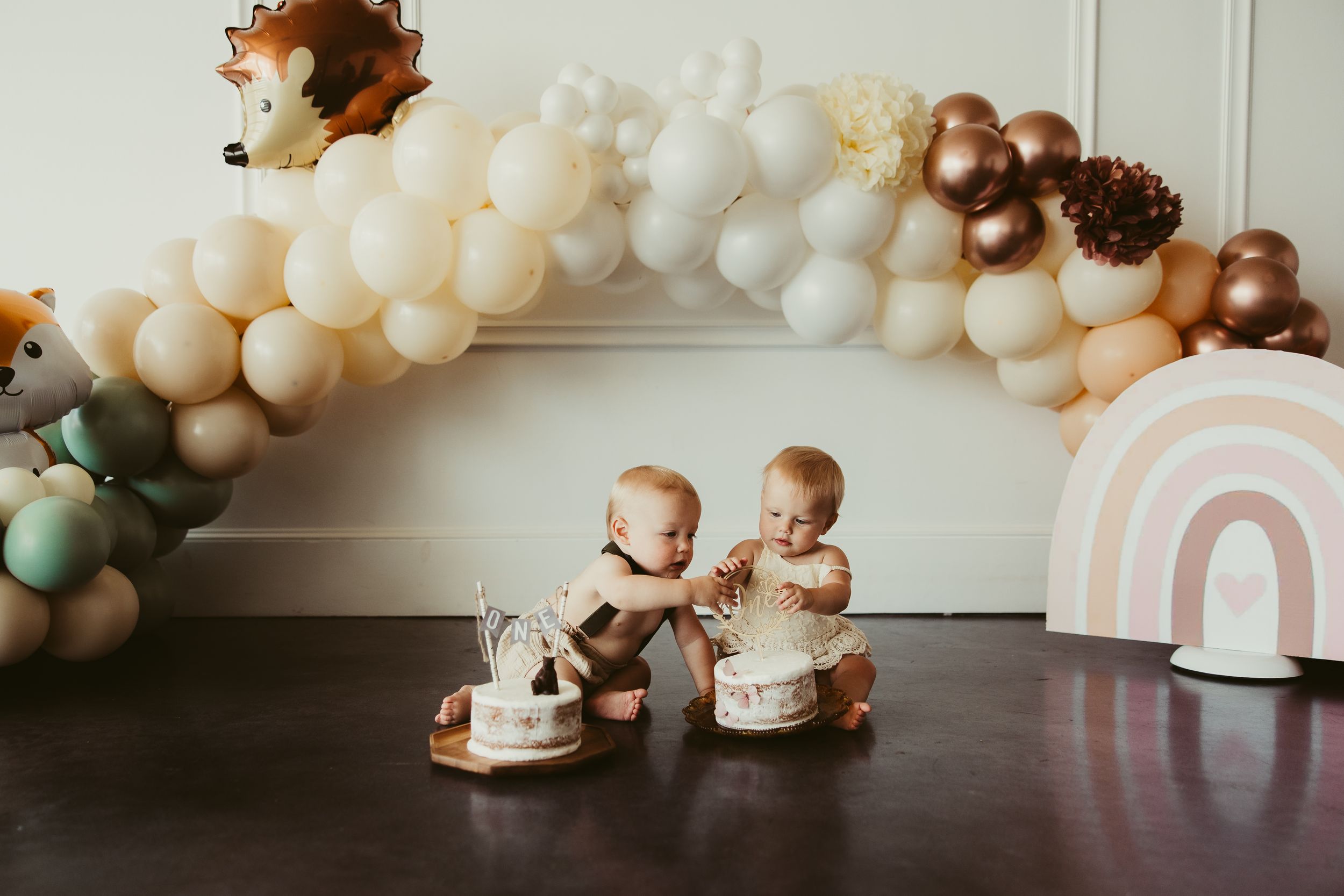 Twin babies celebrate first birthday smashing individual white cakes under a boho woodland balloon garland arch.