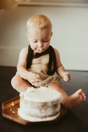 Baby boy in suspenders gleefully smashes his white first birthday cake while sitting on a wooden tray on dark floor.