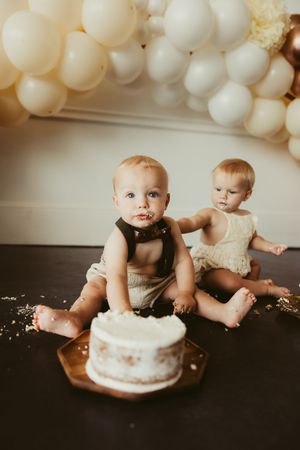 Twin babies sit together with a white smash cake under a cream and white balloon garland at their first birthday.