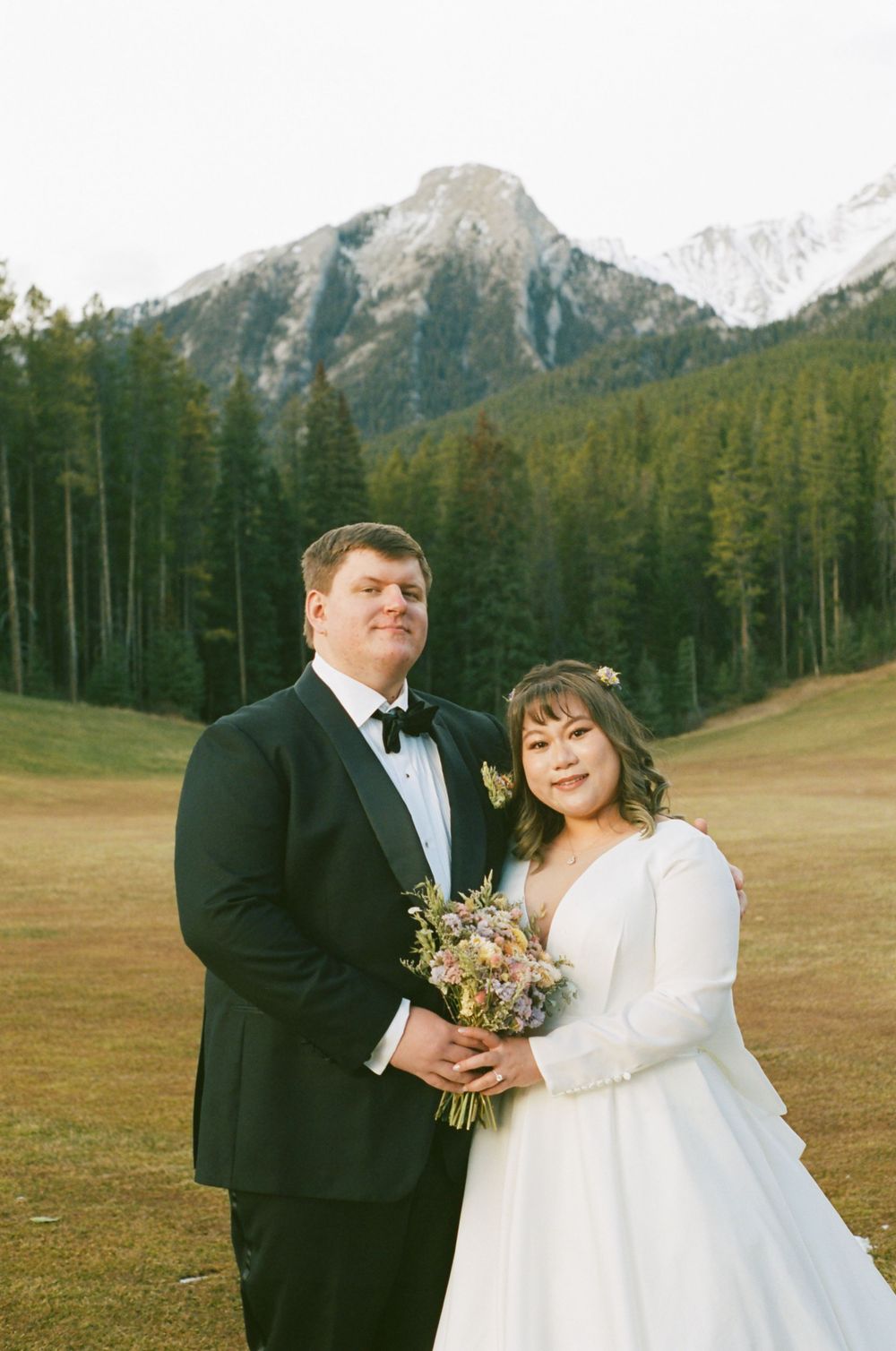 A wedding couple poses in front of majestic mountain peaks and evergreen forest in an outdoor mountain setting.