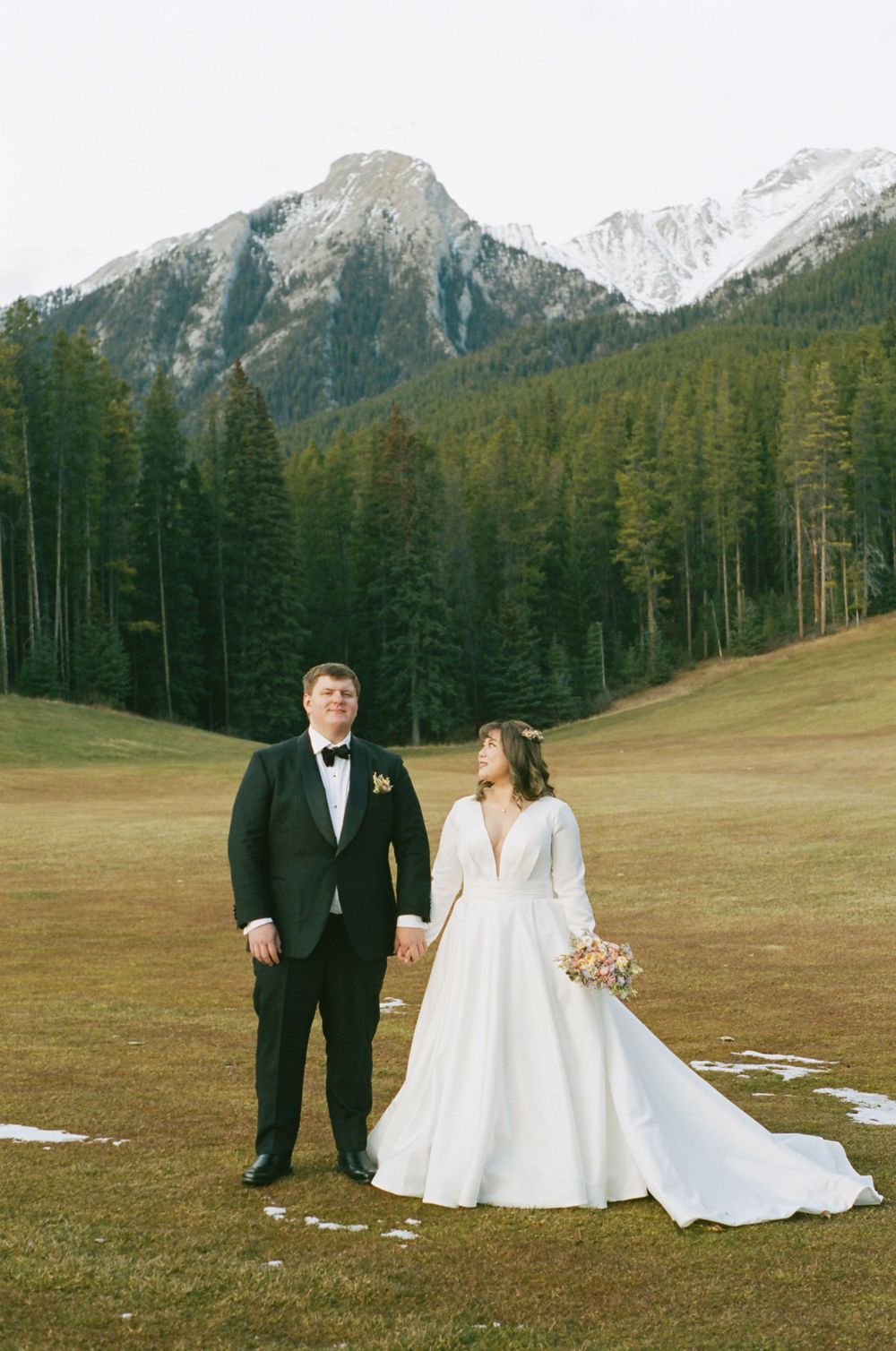 A wedding couple poses in a scenic mountain meadow with snow-capped peaks and evergreen forest in the background.