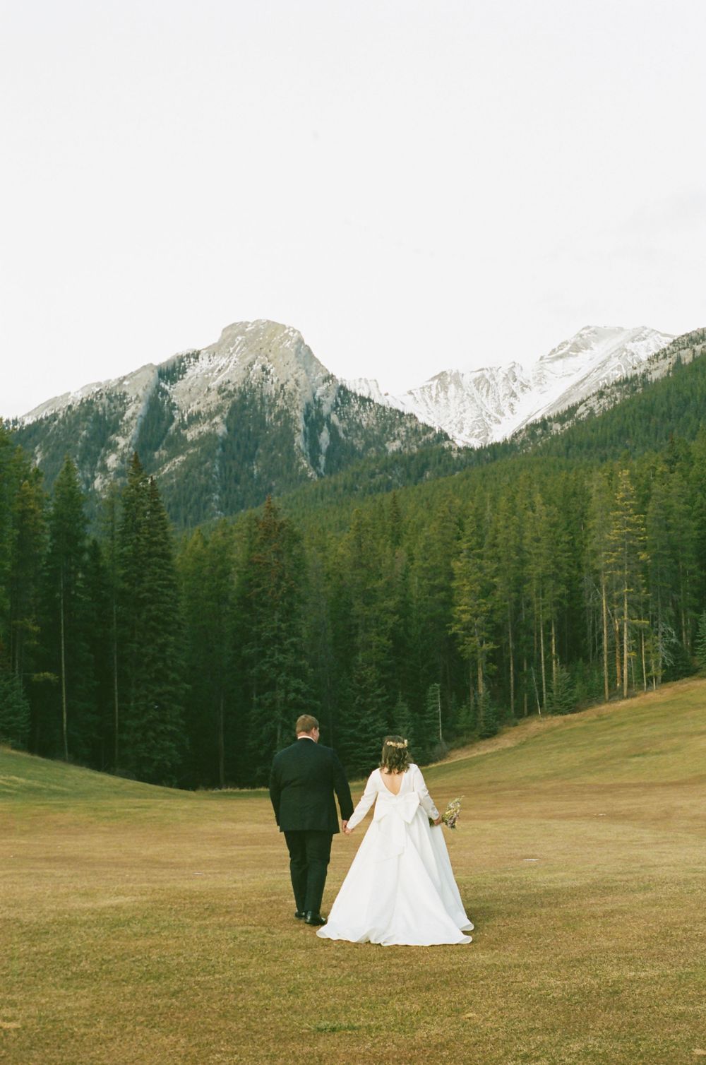 Newlyweds stand together in an alpine field surrounded by majestic mountain peaks and pine forests.