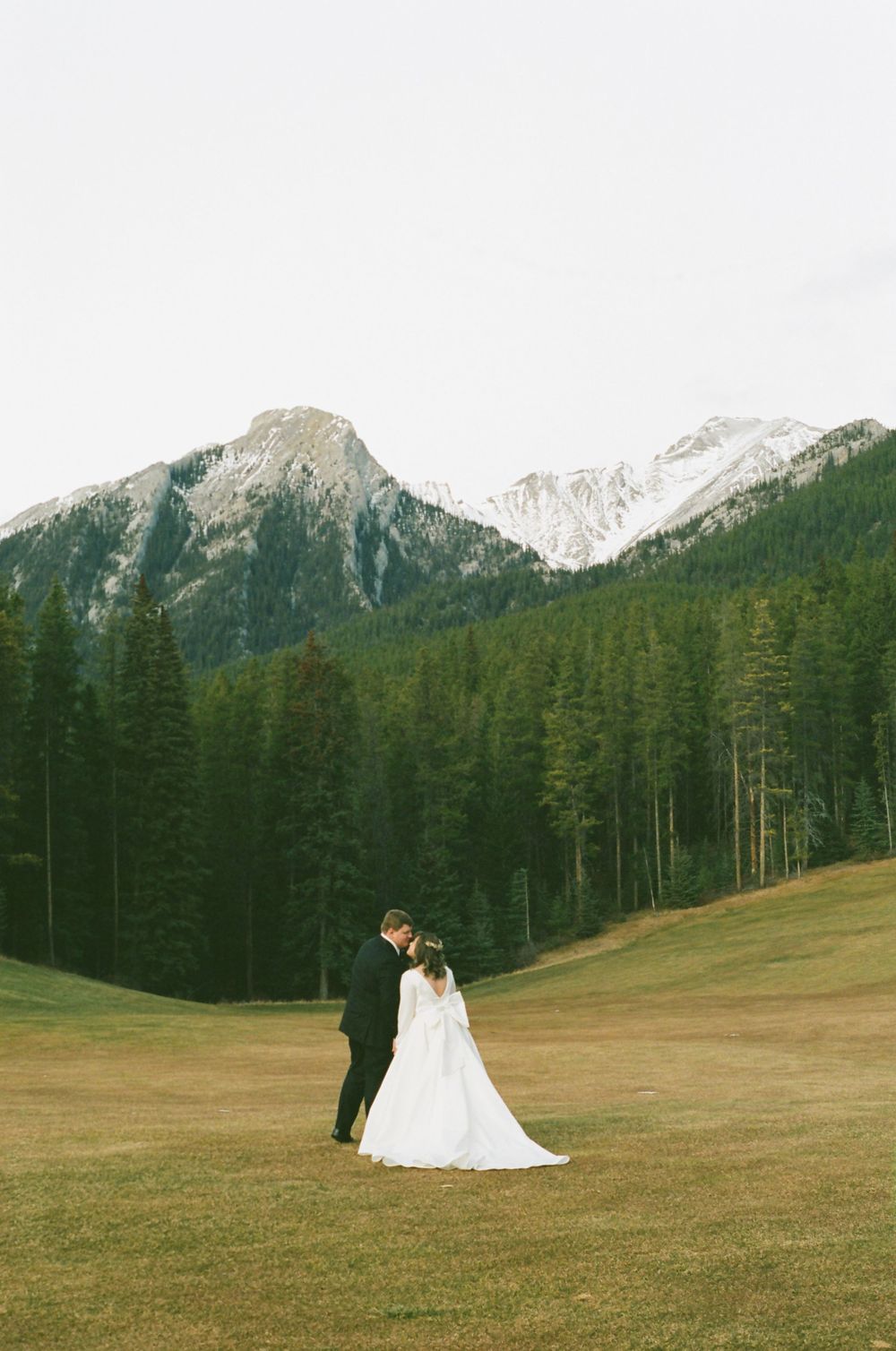 A couple in wedding attire stands in a meadow with majestic snow-capped mountains and evergreen forest in the background.