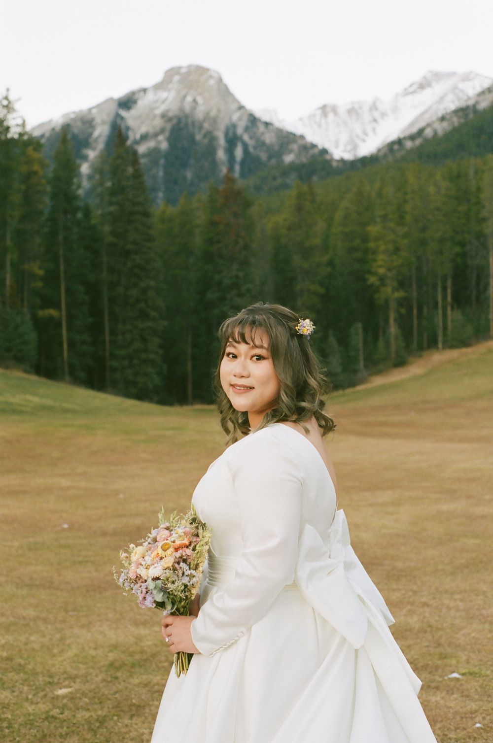 A bride in a white dress with a bow detail holds a bouquet against a mountain landscape backdrop.