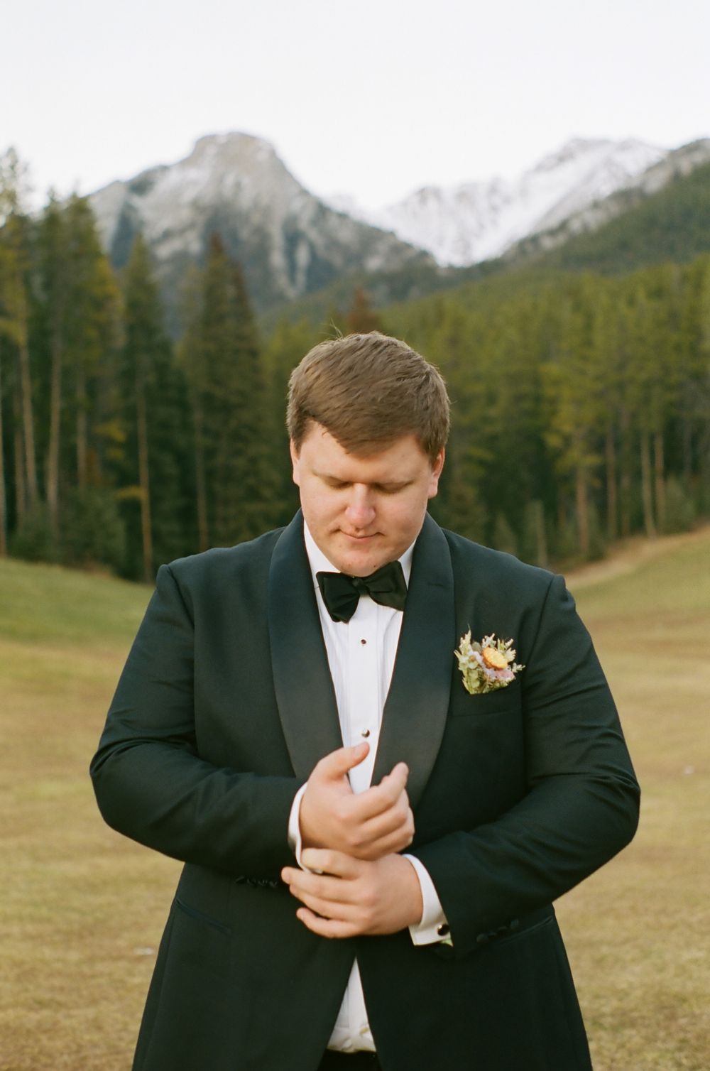 A groom in a black tuxedo adjusts his cuff against a backdrop of snow-capped mountains and pine trees.