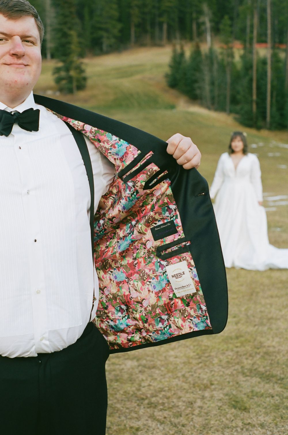 A groom shows off colorful floral lining inside his tuxedo jacket with mountains and forest in background.