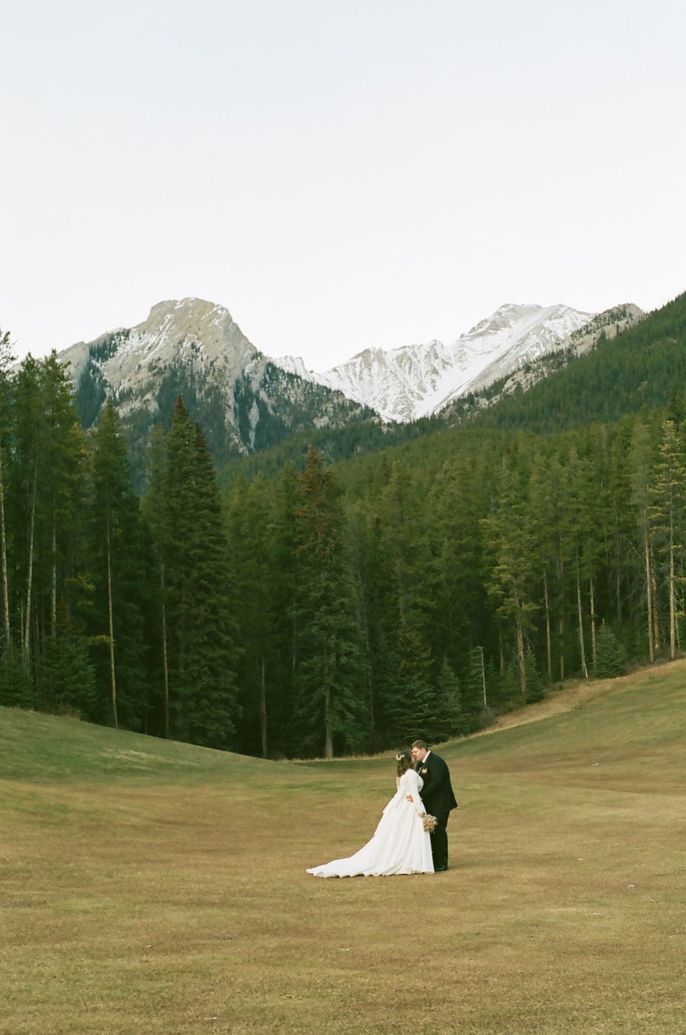 A distant wedding couple stands in a vast mountain meadow with evergreen trees and snow-capped peaks.