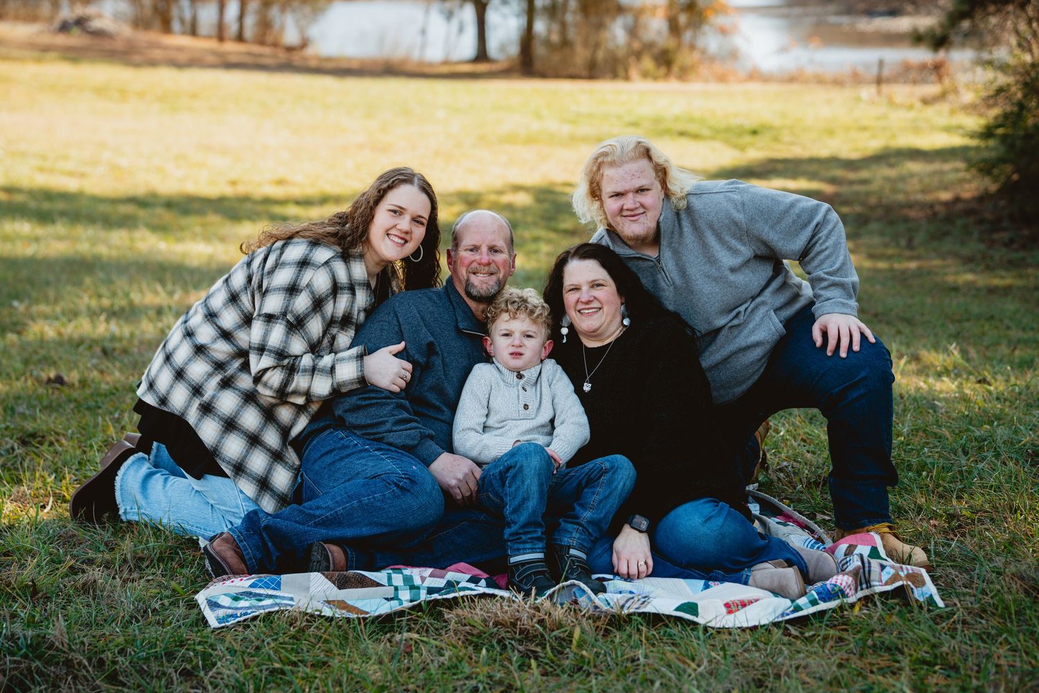 A family group lounges together on a blanket outdoors during an autumn photoshoot in a grassy setting.