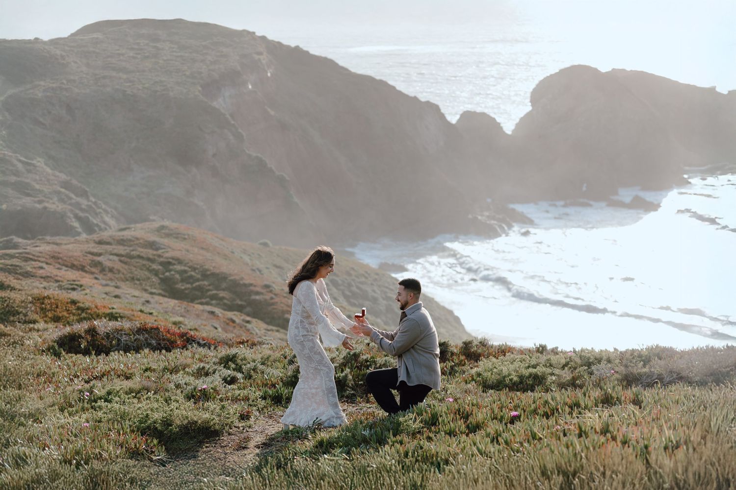 Couple shares romantic moment on scenic coastal cliffside overlooking misty ocean during sunset.