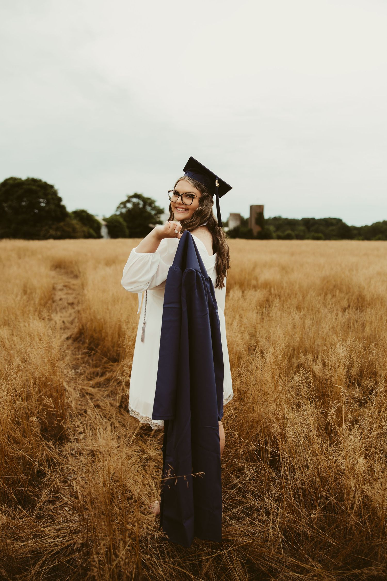 Smiling graduate in cap and gown standing in golden wheat field holding navy blue robe.