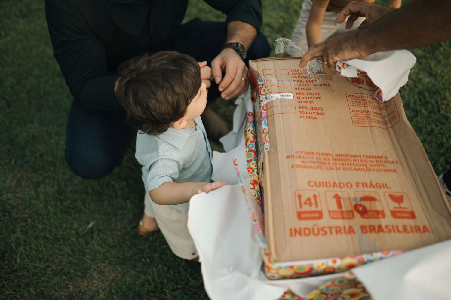 A child in white sits on grass examining the contents of a large cardboard shipping box.