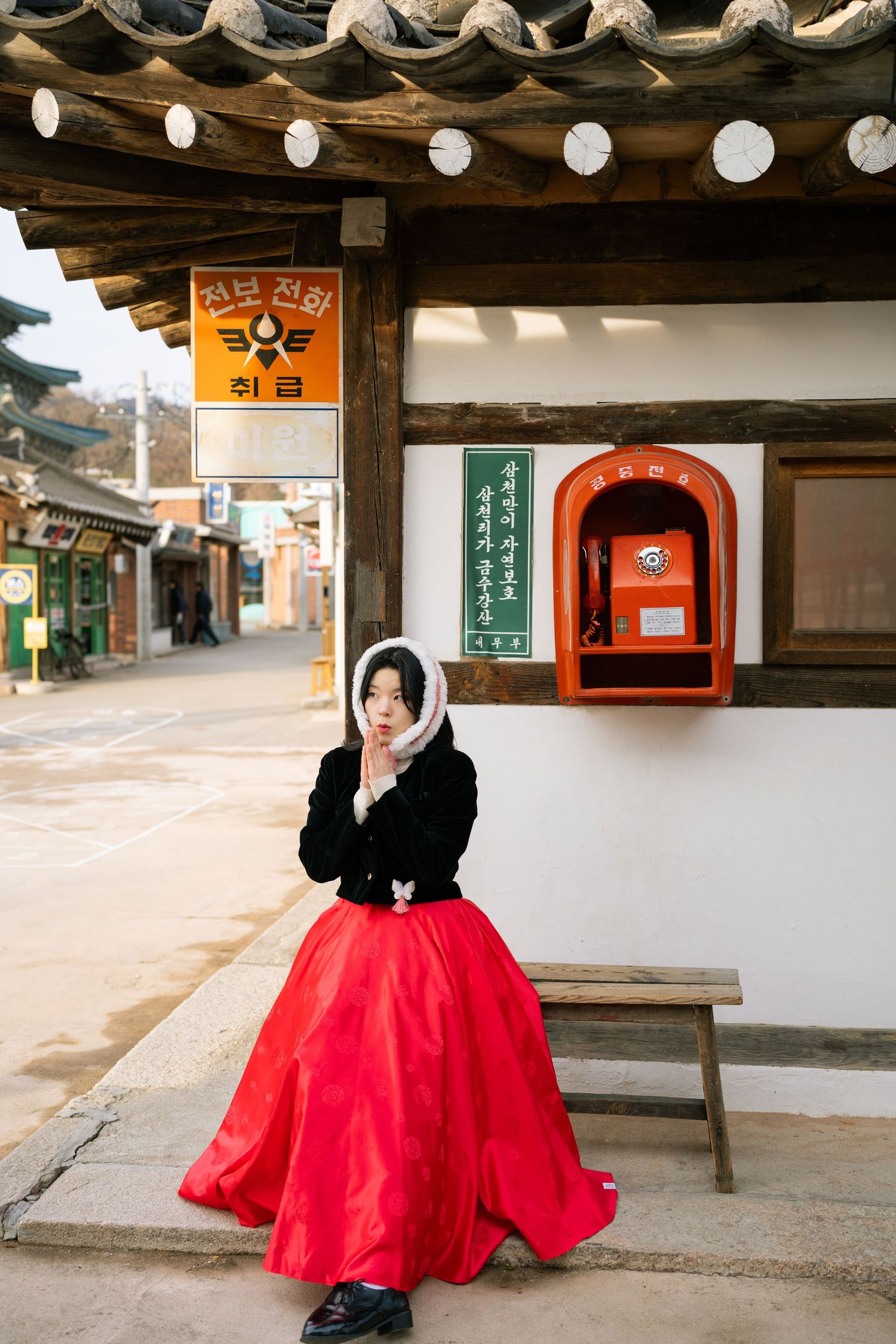 A person in a vibrant red hanbok dress sits on a wooden bench against traditional Korean architecture.