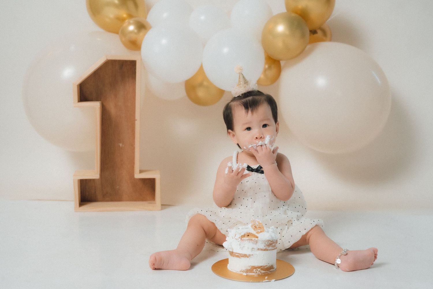 Baby in white dress celebrating first birthday with cake and gold and white balloons.