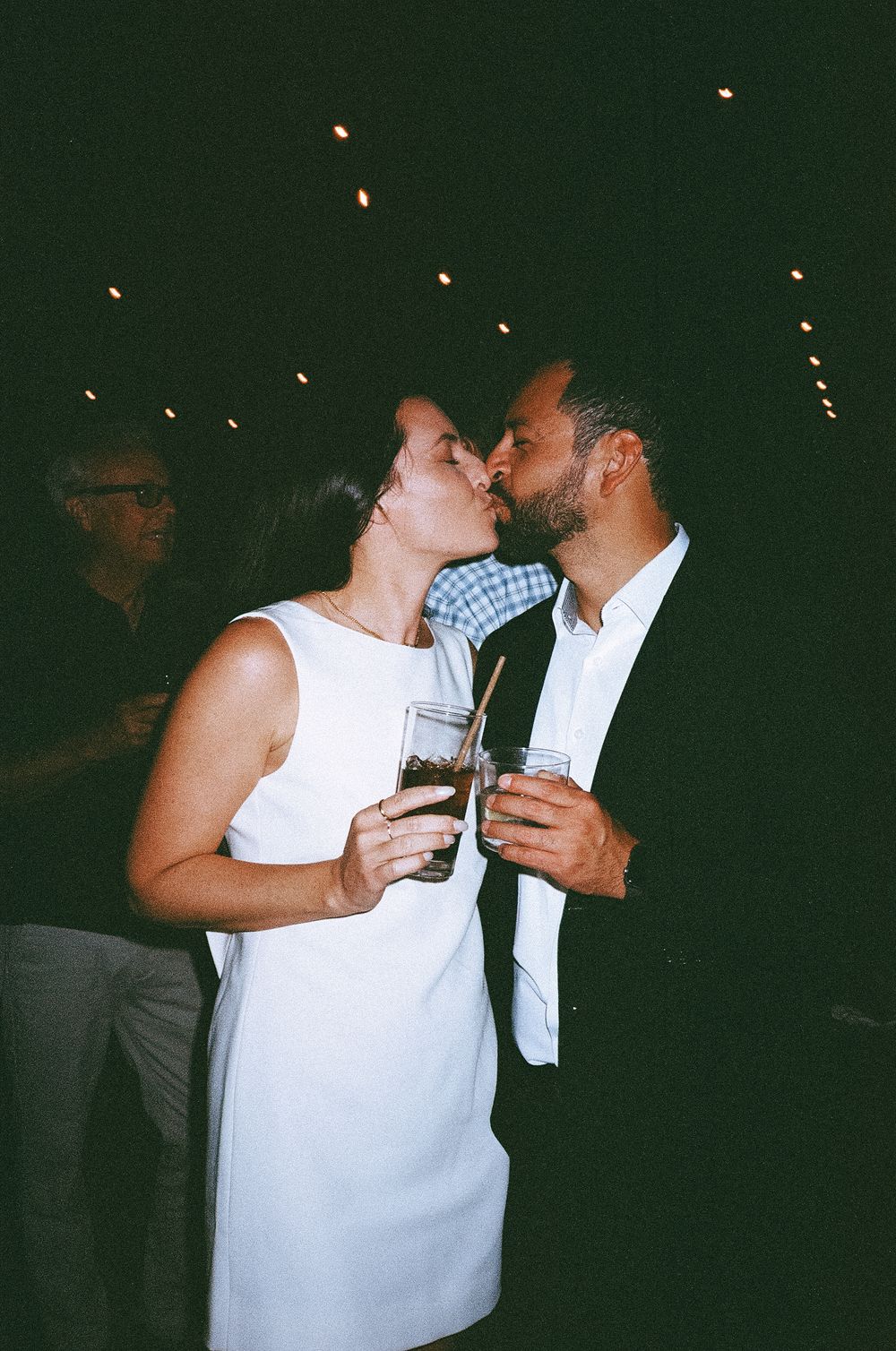 A couple sharing an intimate moment at a dimly lit party or event with twinkling lights in the background.