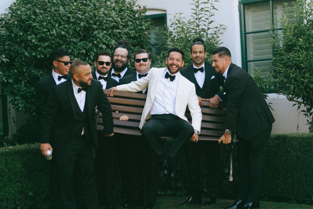 A group of groomsmen in black and white formal attire pose together on a bench outdoors.