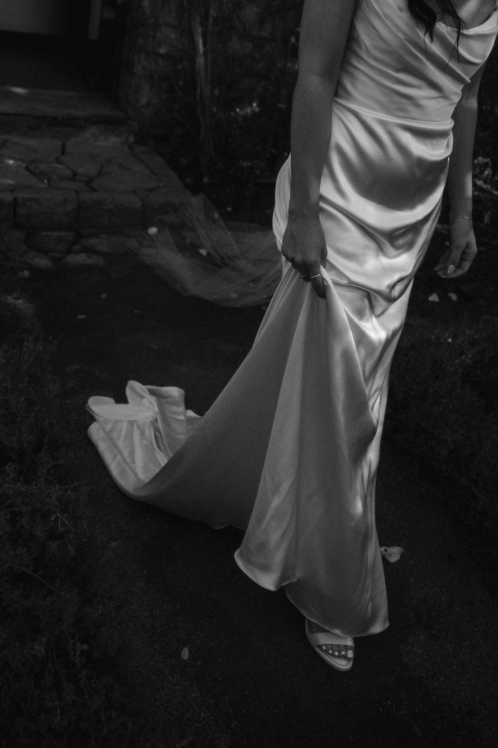 Black and white photo showing the bottom portion of an elegant white satin wedding dress with flowing train and sandals.