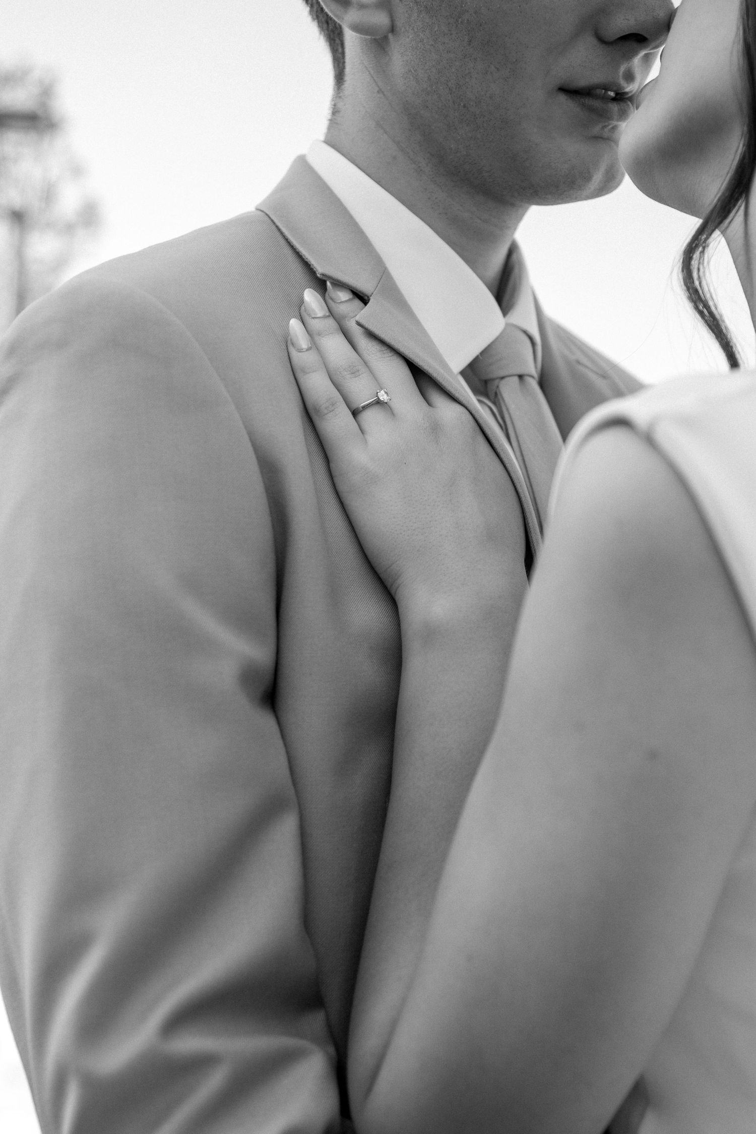 Black and white wedding photo showing a close-up of a groom's suit and tie with a hand wearing an engagement ring.