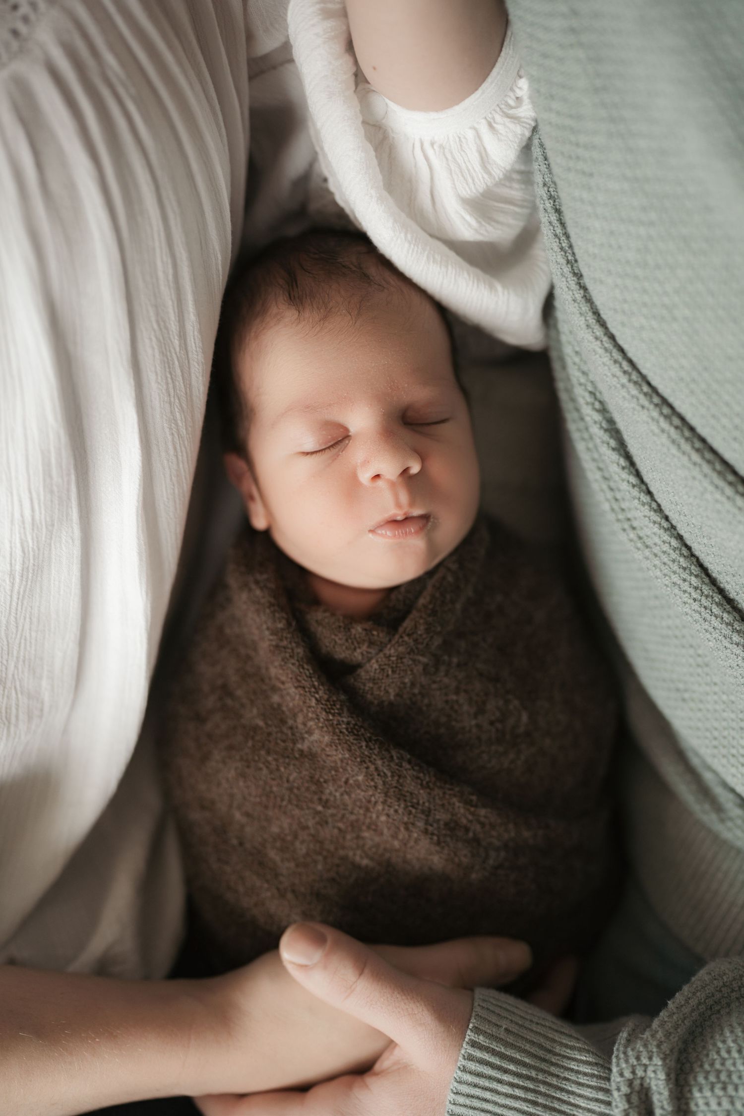 A photo showing a sleeping baby wrapped in a brown blanket with white bedding in soft natural light at a newborn in home session