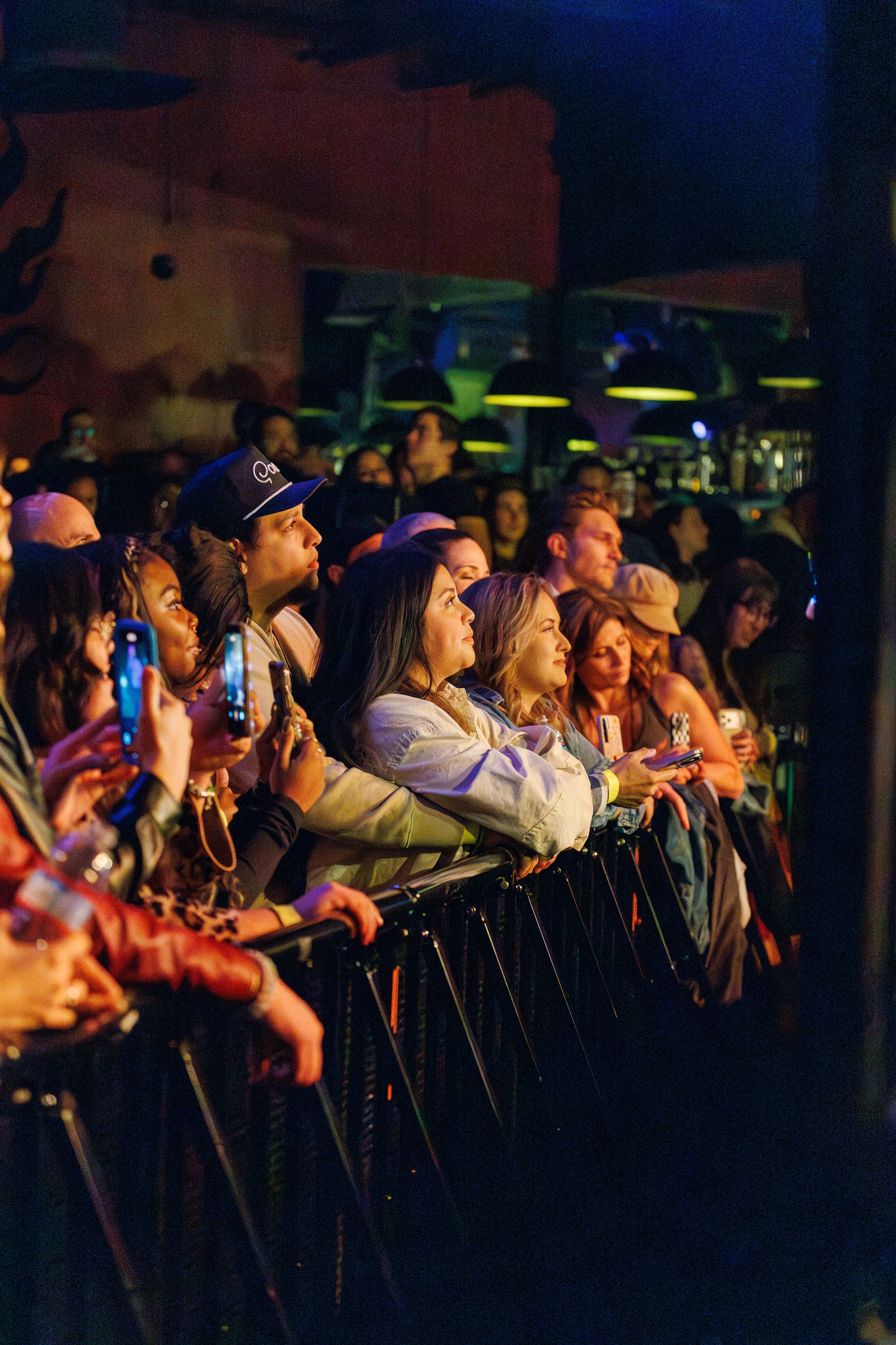 Concert crowd leans against security barrier at night while watching performance in dark venue with colorful lighting.