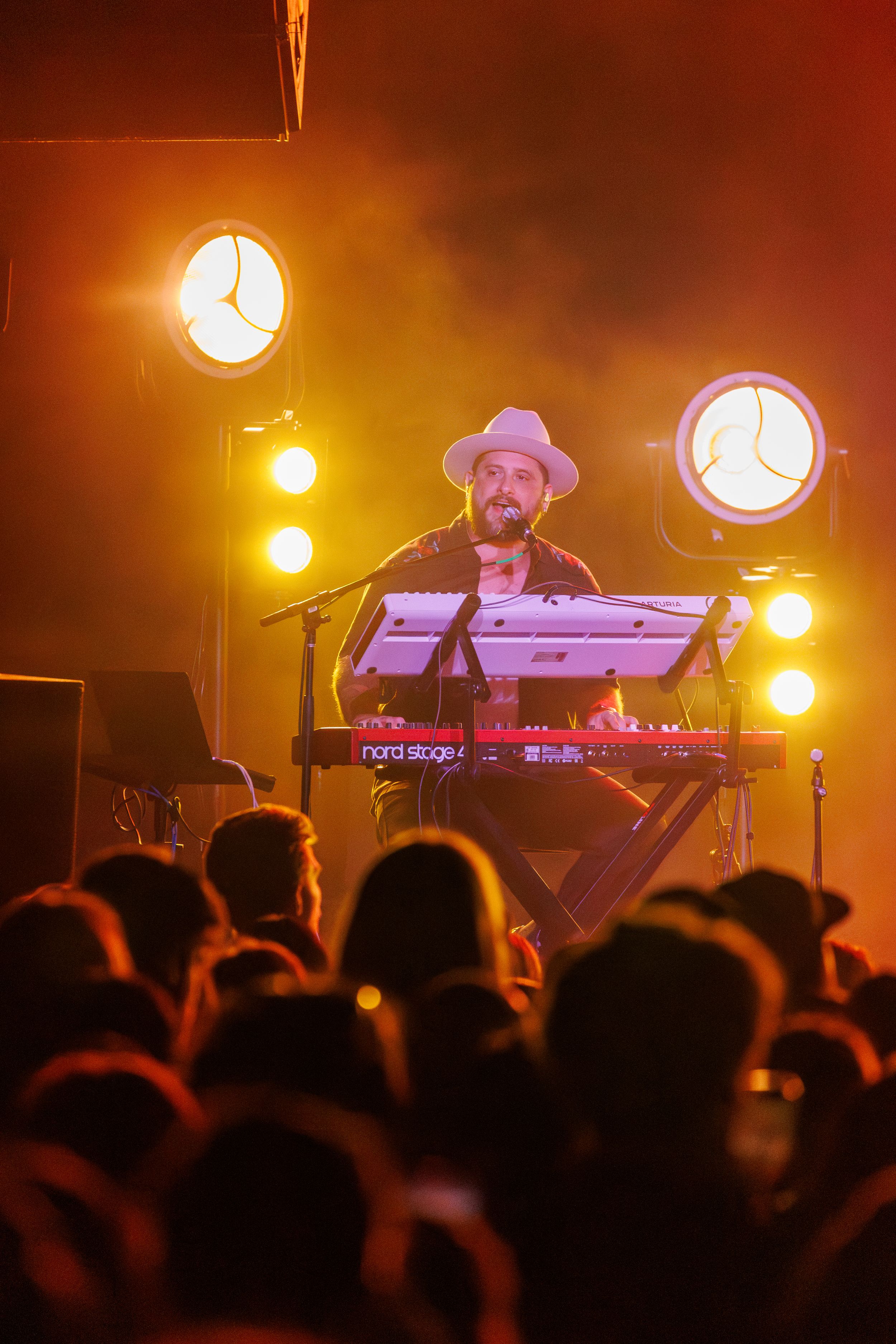 DJ performs on stage with electronic equipment under warm orange lighting as crowd silhouettes dance below.