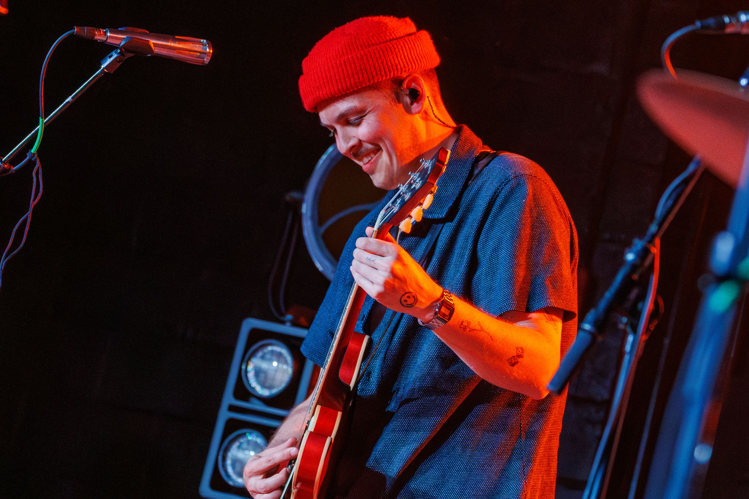 Musician in red beanie and blue shirt plays guitar on stage under dramatic red lighting during live performance.