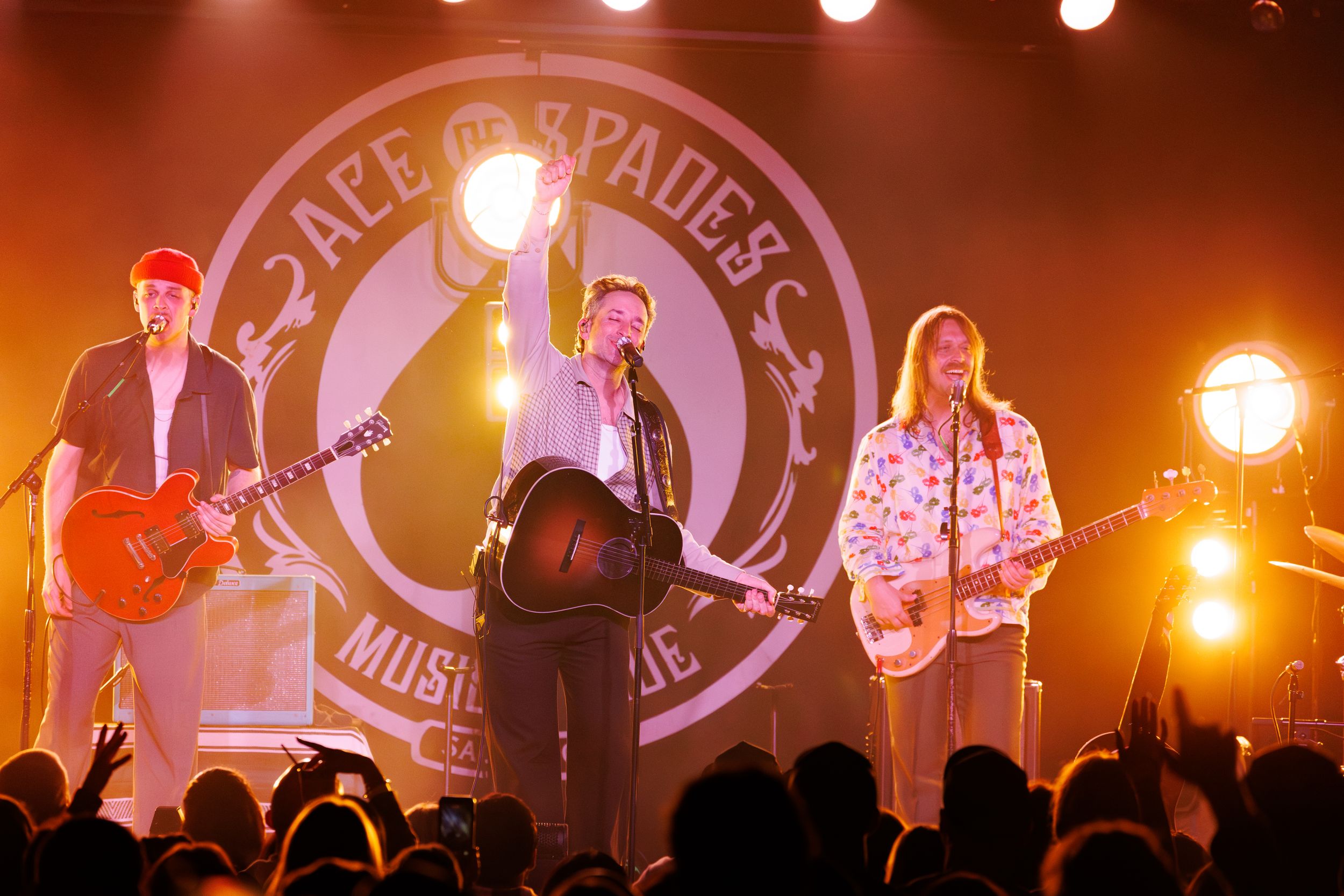 Musicians perform on stage at a concert with warm orange lighting and a large illuminated logo backdrop.
