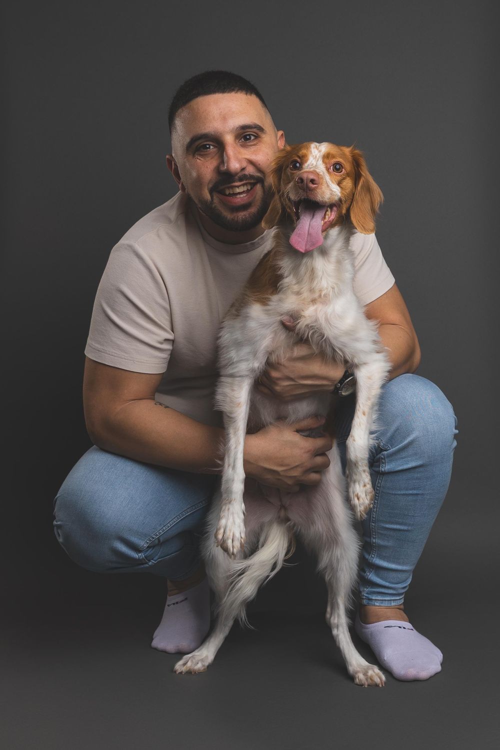 Man in white t-shirt and jeans crouching while holding a happy Brittany Spaniel dog against a gray background.