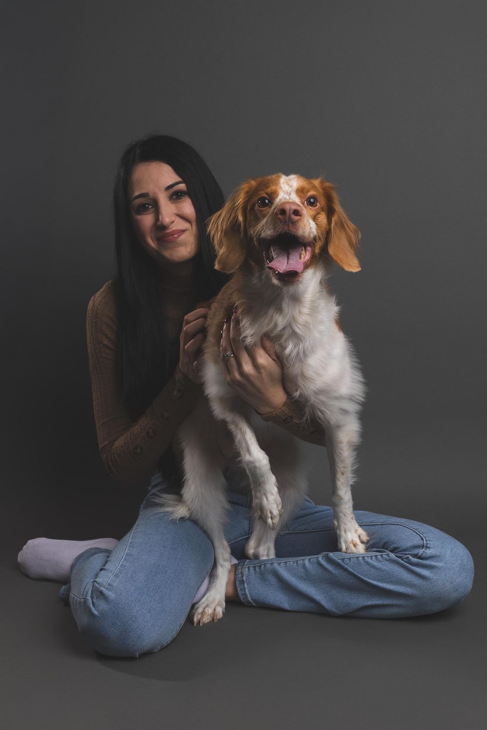 Young woman with dark hair sits cross-legged on gray background, smiling while holding a happy Brittany Spaniel dog.