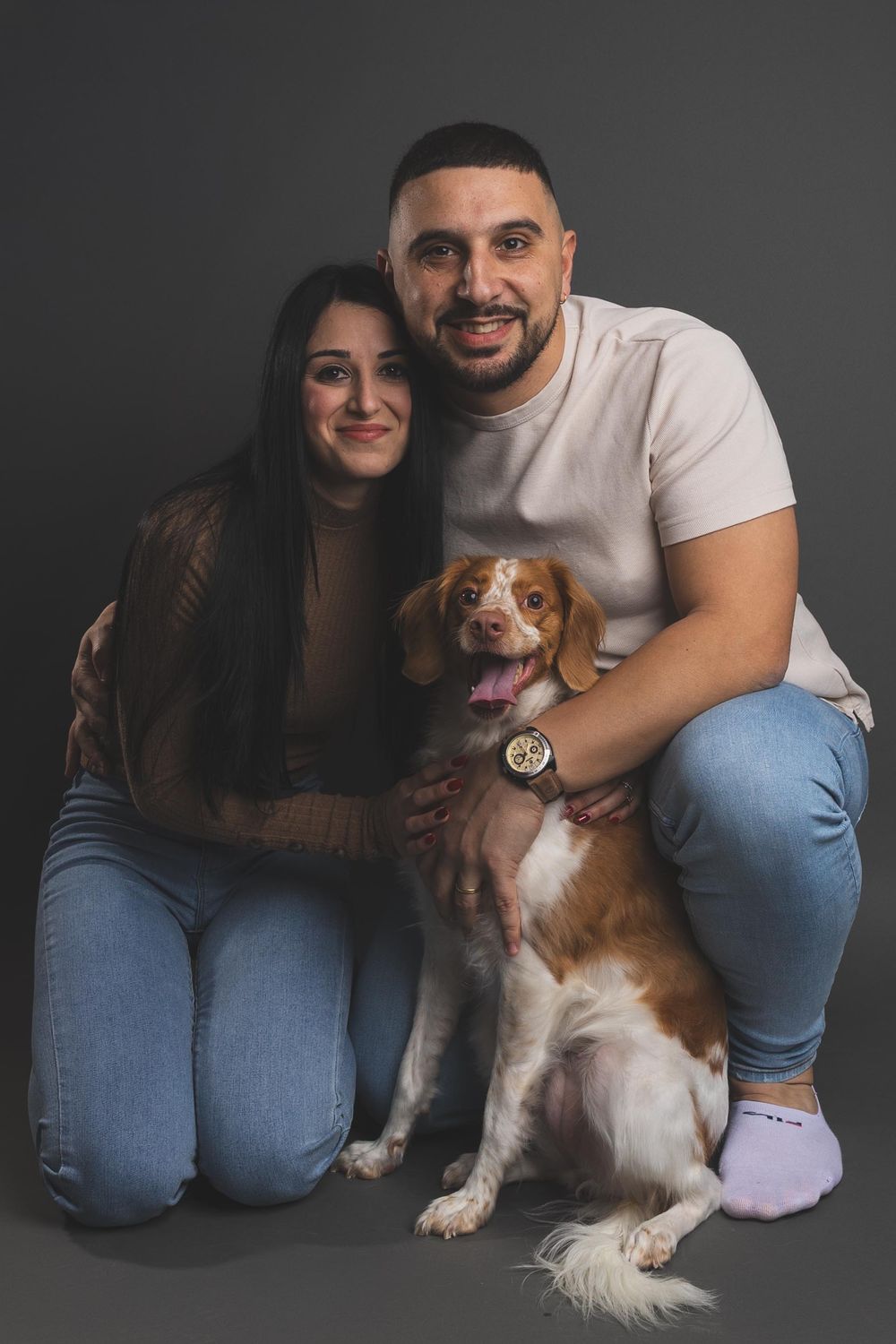 Smiling couple in casual clothes poses with their brown and white Brittany Spaniel dog.
