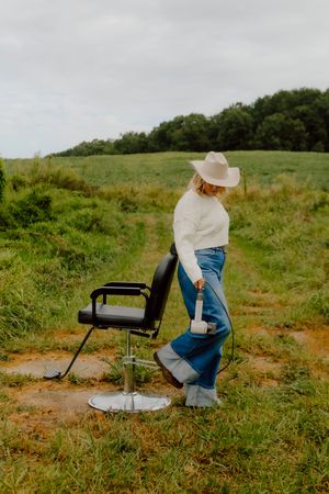 Woman in white sweater, jeans, and cowboy hat standing beside barber chair in field.