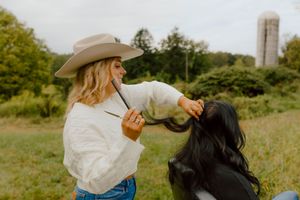 Woman in cowboy hat playfully braiding another woman's long black hair in outdoor field.