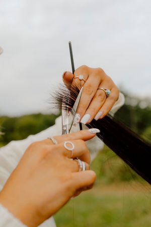 Close-up of hands with rings cutting dark hair with scissors outdoors.